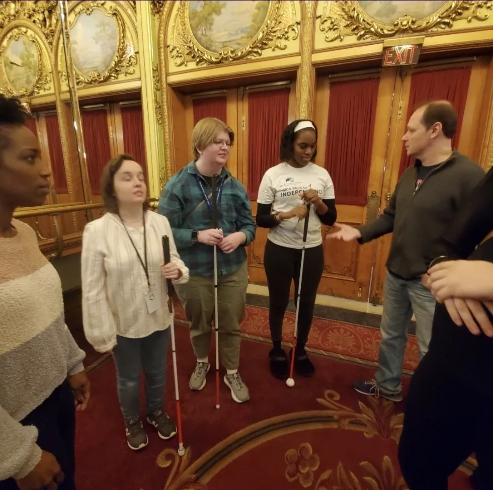 Students from the Carroll School for the Blind stand with their canes,
            listening to Into the Woods cast members Jim Stanek and Felicia Curry talk
            about some of the props and costumes during a guided touch tour in
            Boston, MA 2023.