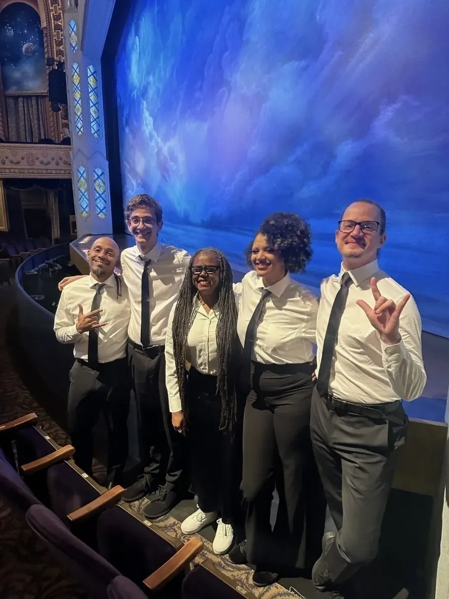 A group of ASL interpreters stand smiling with their arms around one
        another in front of the stage of Book of Mormon. L-R; Alberto Medero, Mark Weissglass, Patrice Creamer, Osaze Ogbeide,
        Patrick Michael