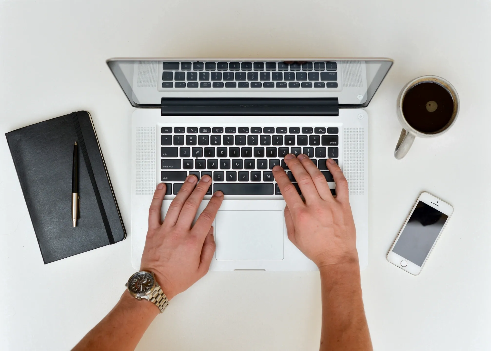 A birds eye view of hands typing on a laptop. A black notebook and
            pen are to the left of the laptop and to the right is a cup of black coffee
            and a white Iphone.